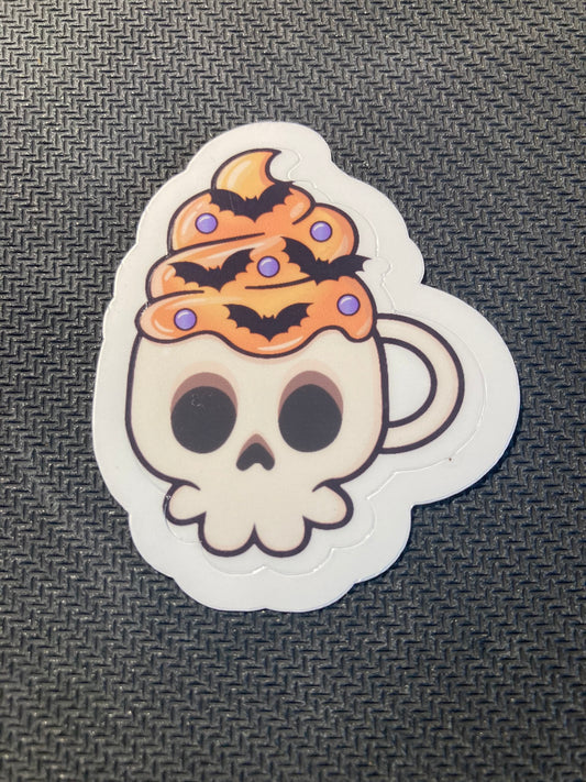 Halloween sticker of a skull-shaped coffee mug filled with orange whipped topping, bats, and sprinkles.