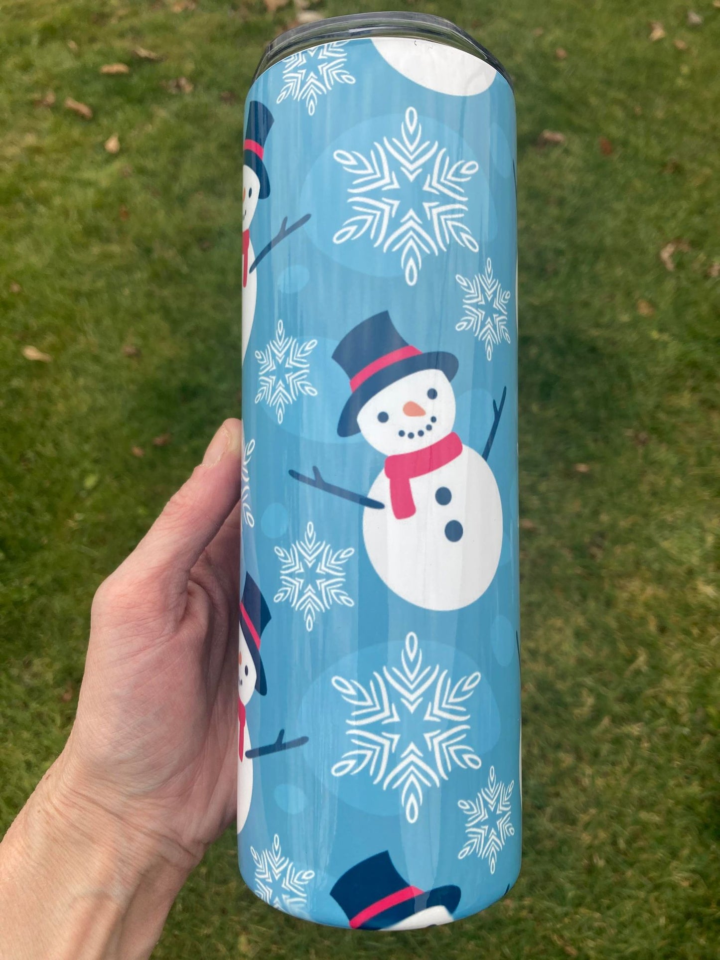 Hand holding a 20oz snowman Christmas tumbler with blue snowflake background outdoors.