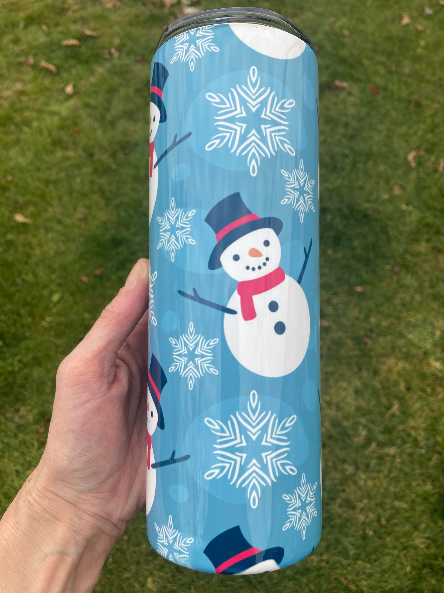 Hand holding a 20oz snowman Christmas tumbler with blue snowflake background outdoors.