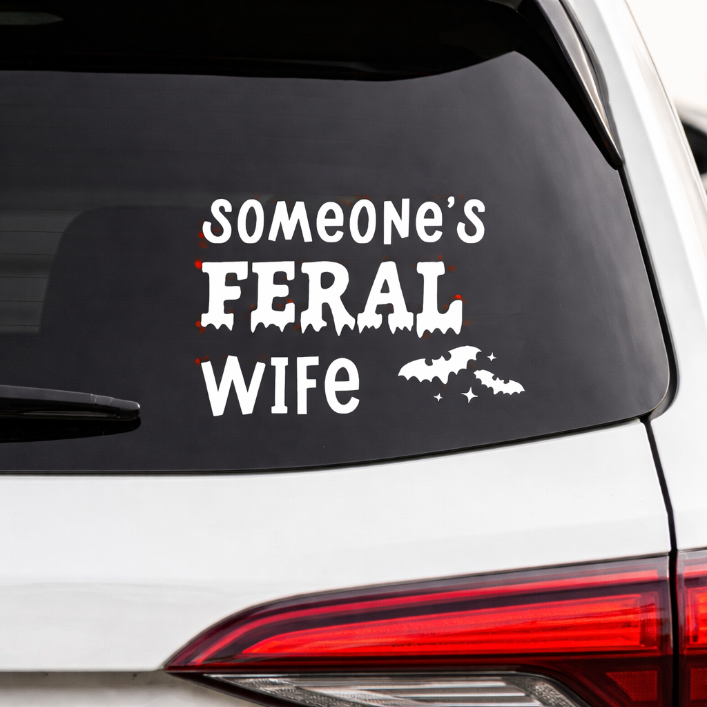 Car window with a decal reading 'Someone's Feral Wife' on a white vehicle.