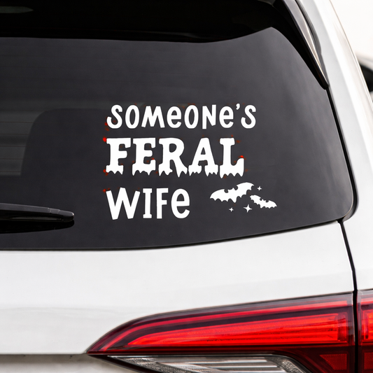 Car window with a decal reading 'Someone's Feral Wife' on a white vehicle.