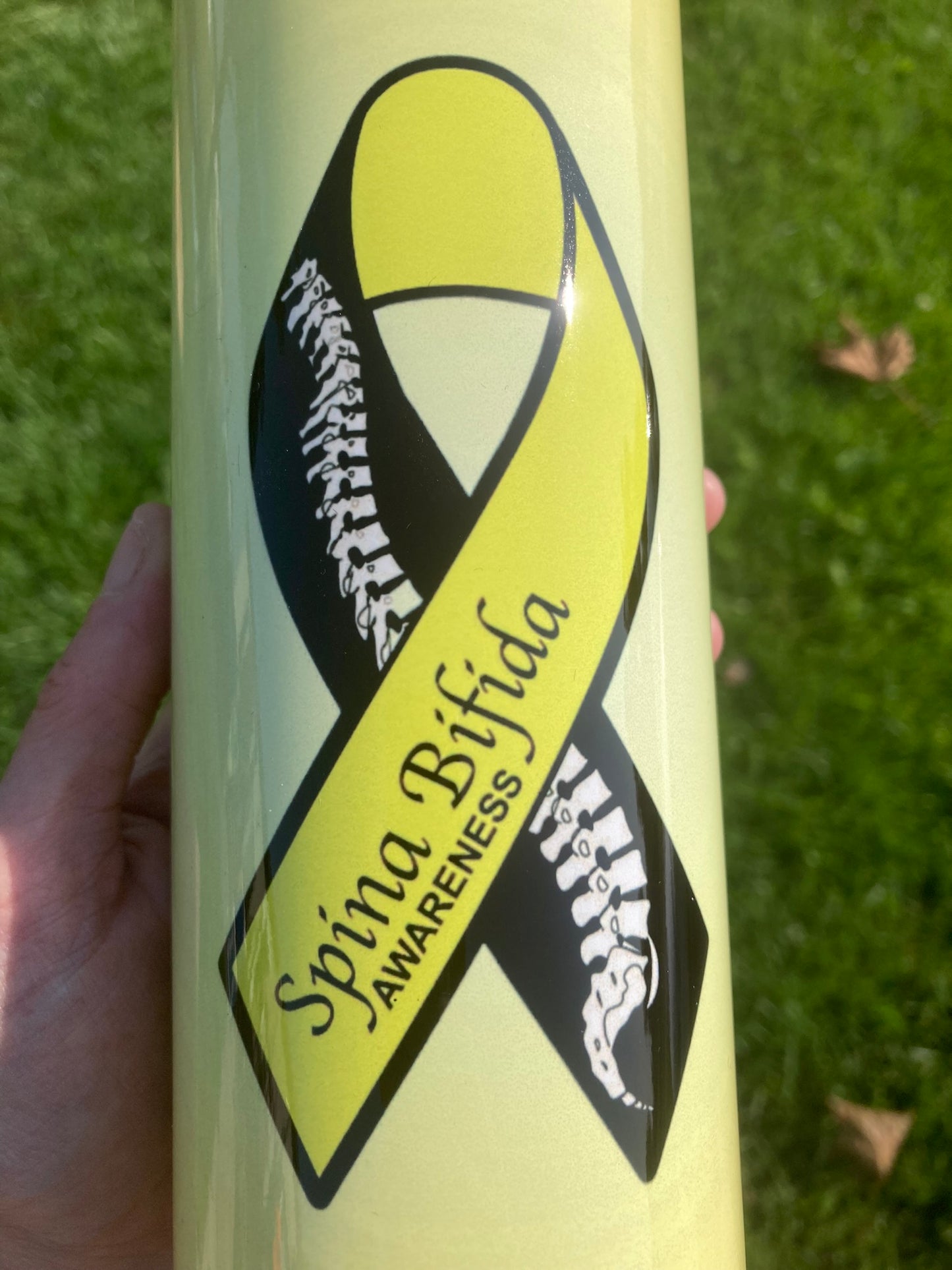 Tumbler with Spina Bifida awareness ribbon design held against a grass background