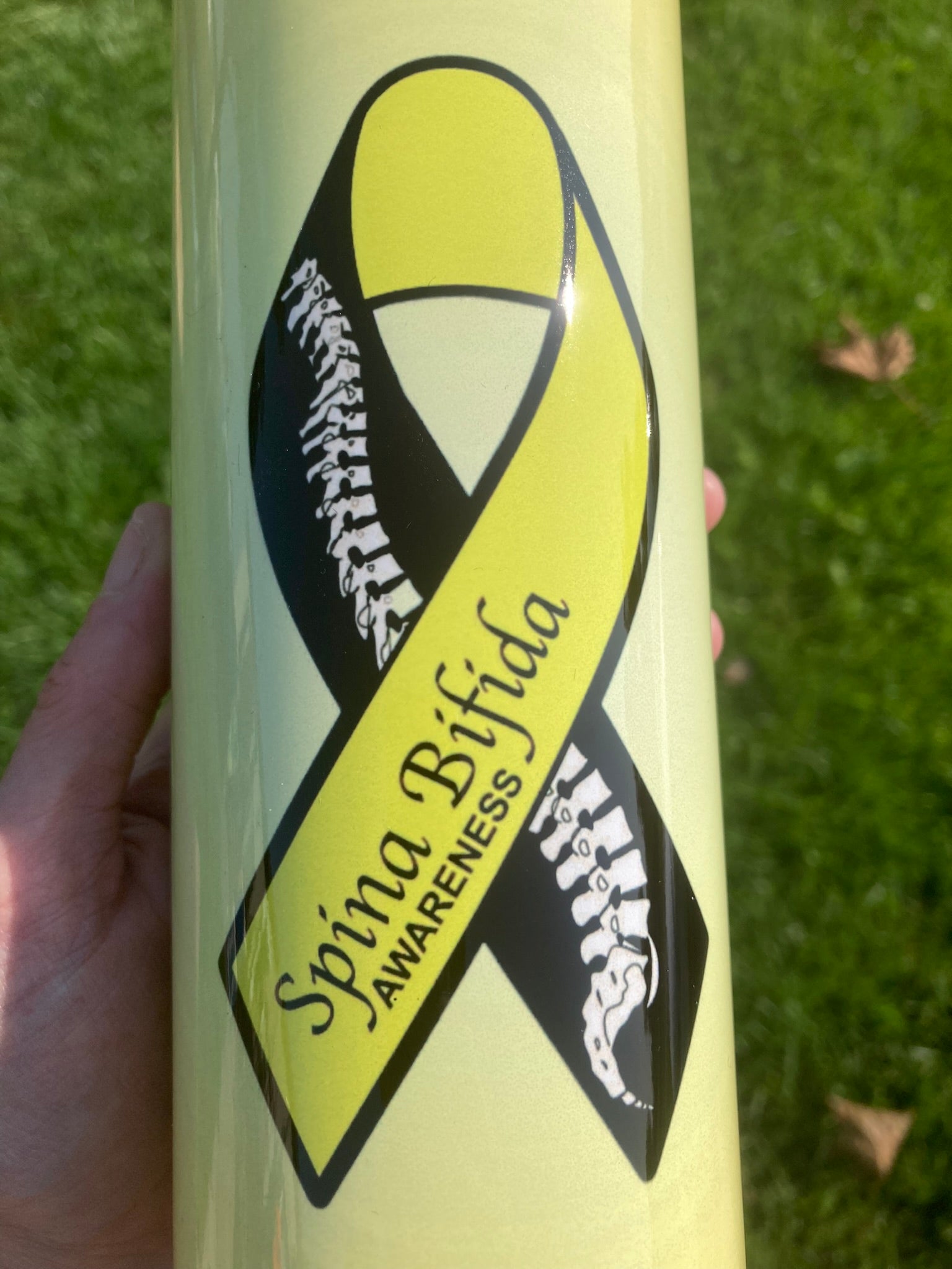 Tumbler with Spina Bifida awareness ribbon design held against a grass background