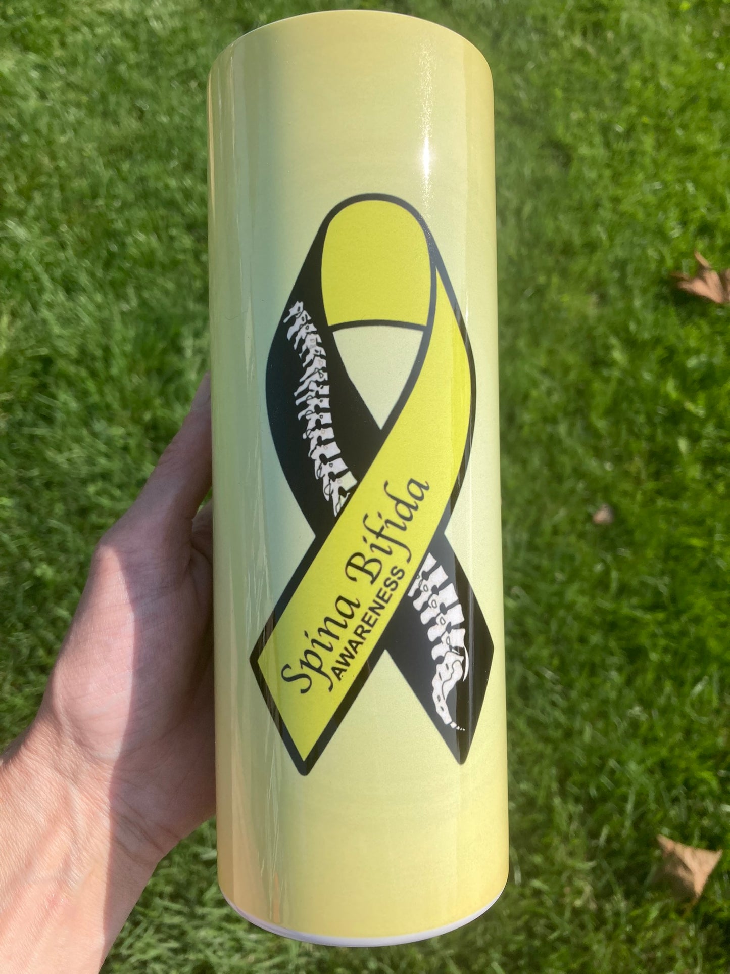 Yellow tumbler with Spina Bifida awareness ribbon held in a hand on grass