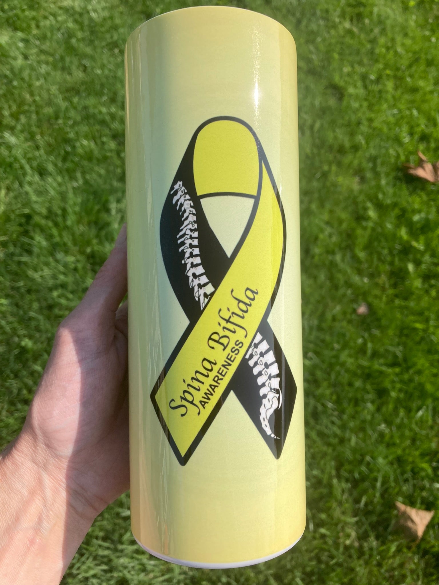 Yellow tumbler with Spina Bifida awareness ribbon held in a hand on grass
