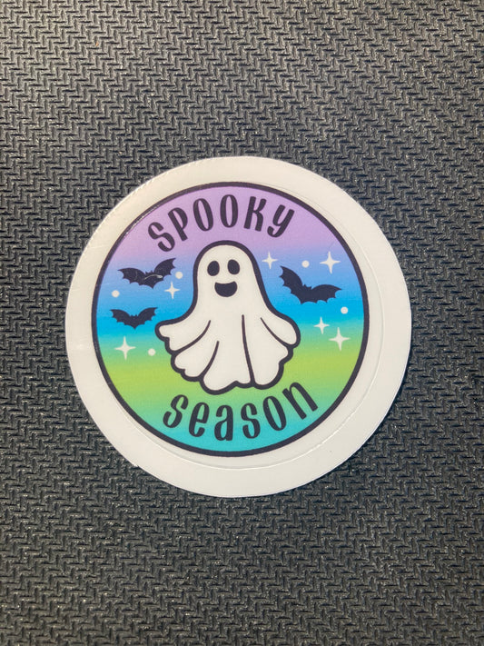 Round Halloween sticker with smiling ghost, bats, sparkles, and rainbow pastel background with text “Spooky Season.”