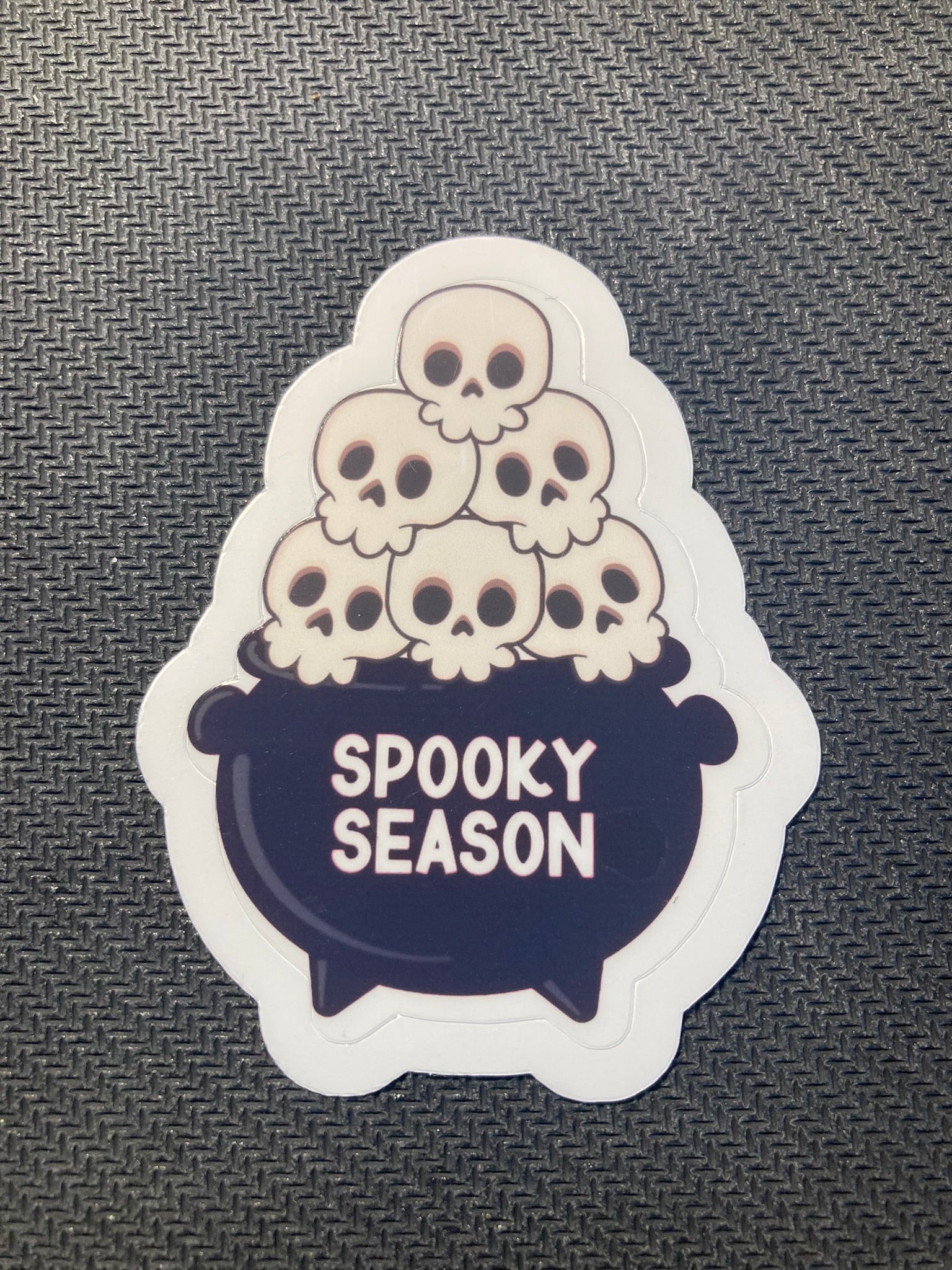 Halloween sticker with a black cauldron filled with cartoon skulls and the words “Spooky Season.”