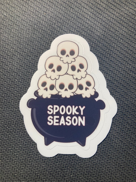 Halloween sticker with a black cauldron filled with cartoon skulls and the words “Spooky Season.”