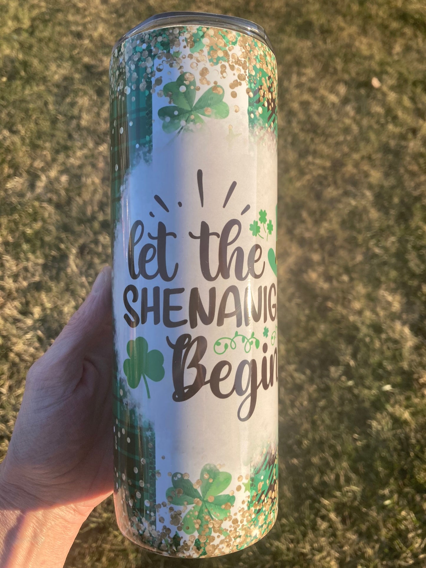 Tumbler with 'let the shenanigans begin' text and shamrock design held by a hand on a textured surface.
