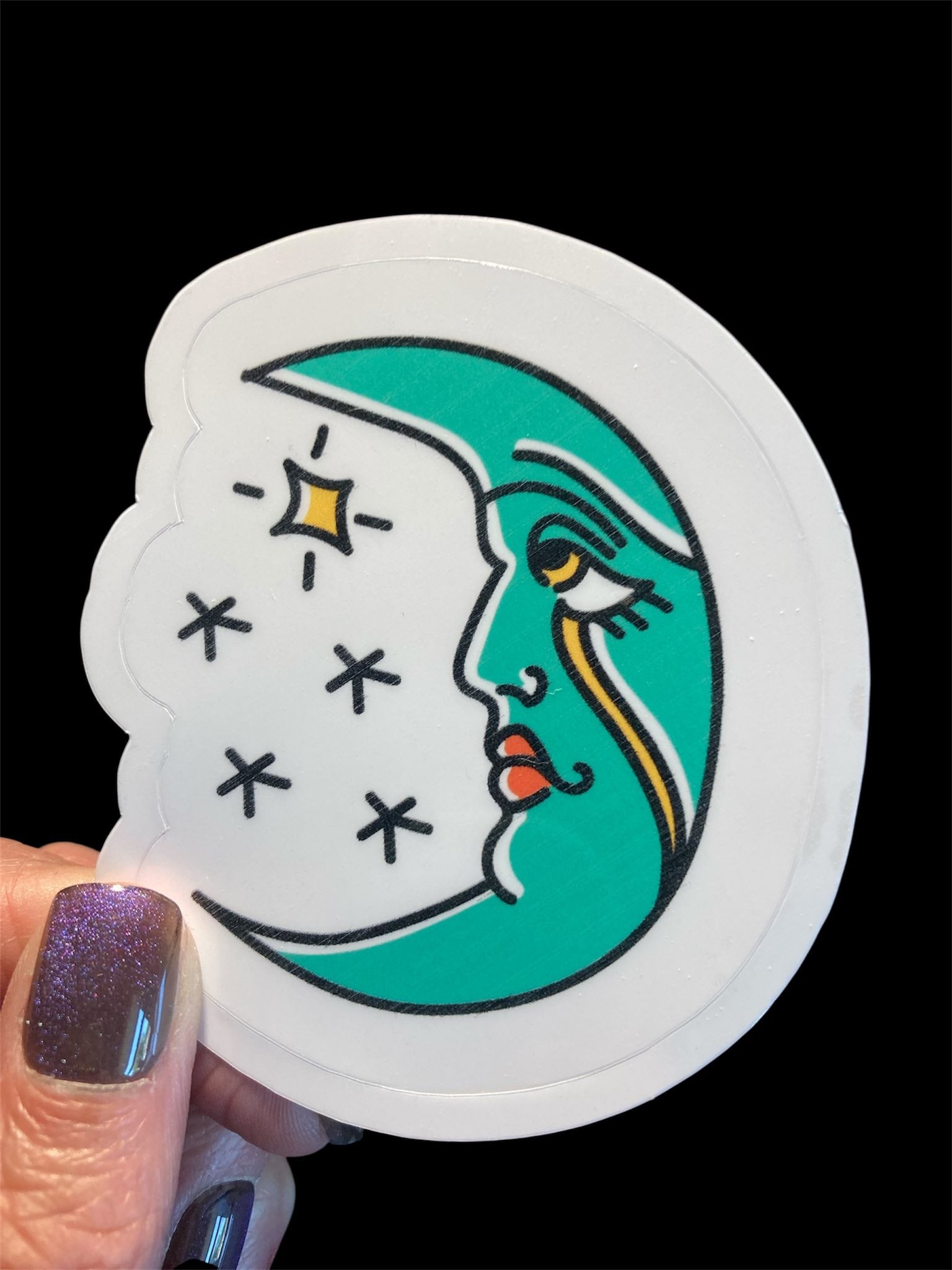 Sticker with a design of a face on a crescent moon with stars, held by a hand.