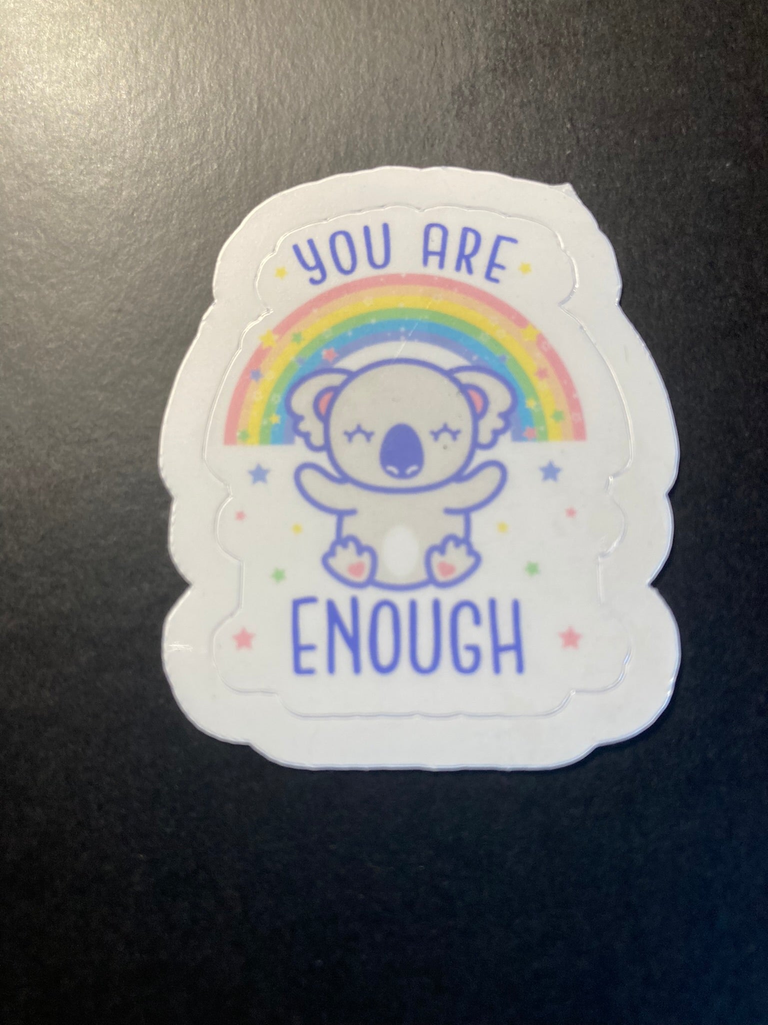 Sticker with a bear illustration, rainbow, and 'You are enough' text on a black surface.
